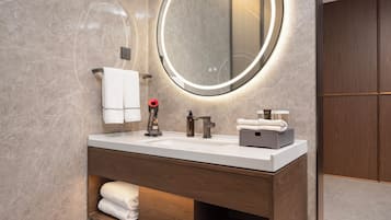 A modern bathroom with a marble wall, a round mirror with underglow lighting, a wooden vanity, and a towel rack.