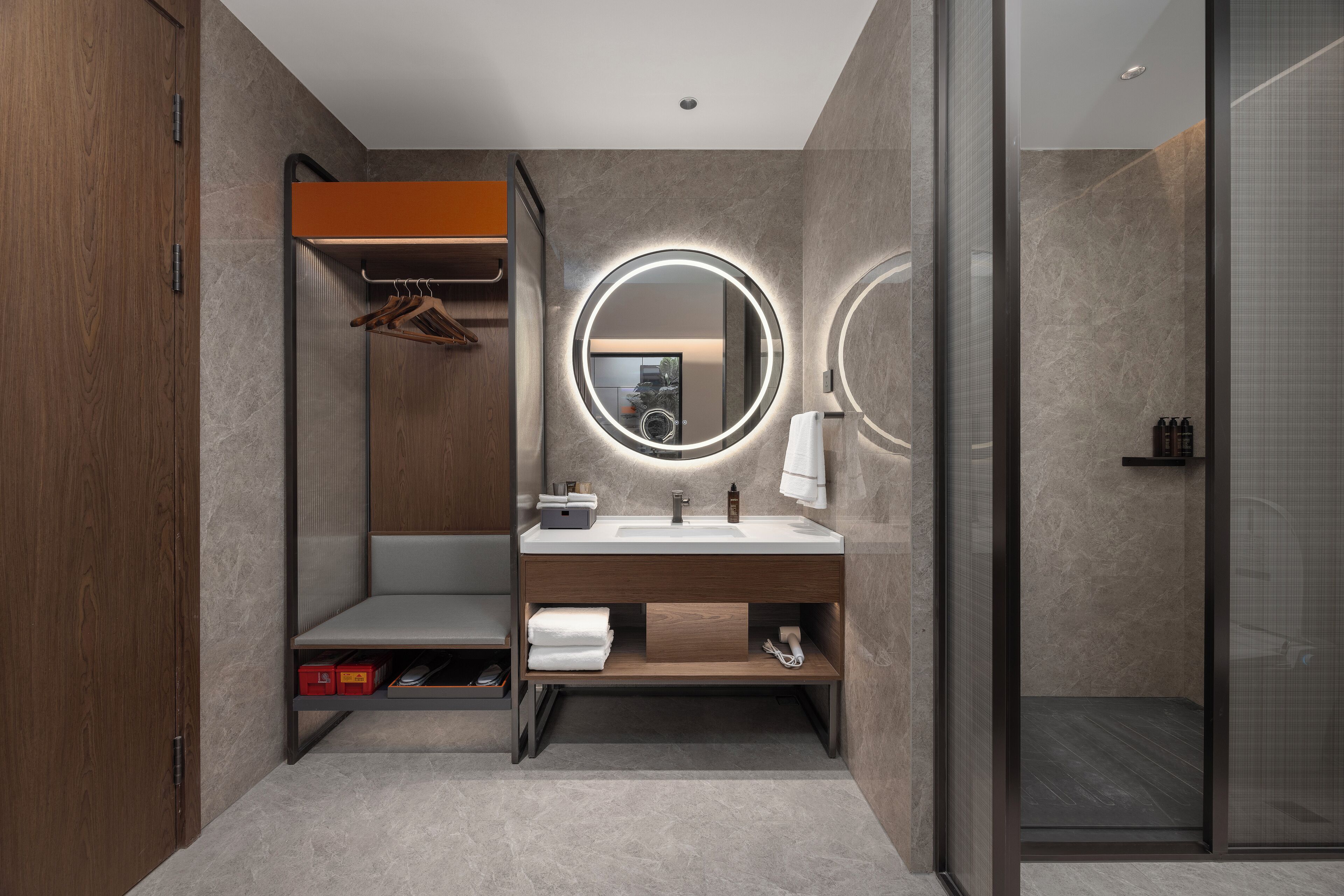 Un baño moderno con tocador de madera, un espejo redondo con iluminación indirecta y cabina de ducha de vidrio, además de un banco empotrado.