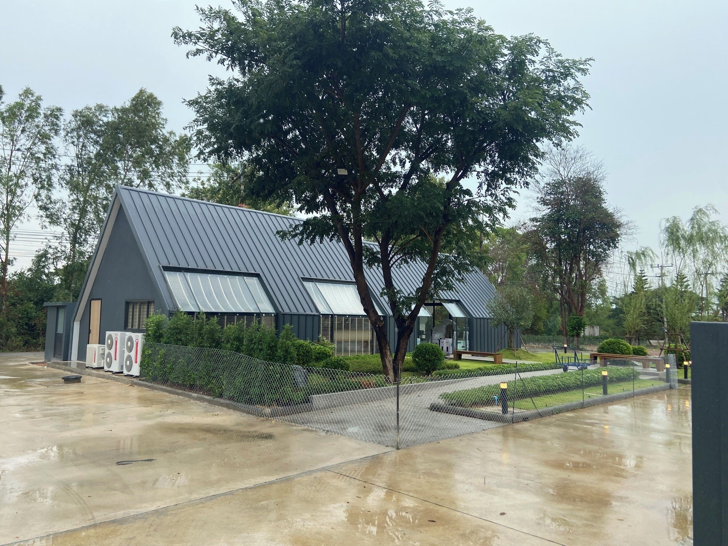 Un edificio moderno con techo inclinado, ventanas amplias y un área pavimentada con valla.