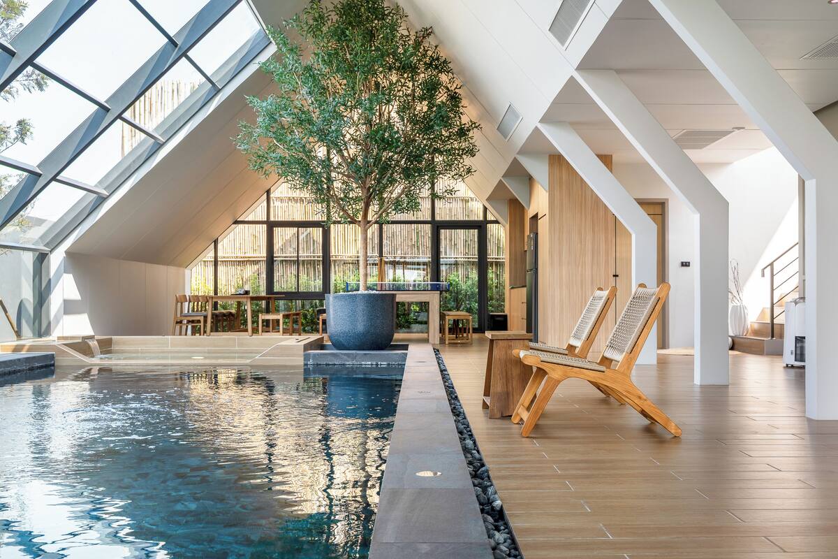 An indoor swimming pool with a wooden floor, a large potted tree, and a seating area with wooden chairs.
