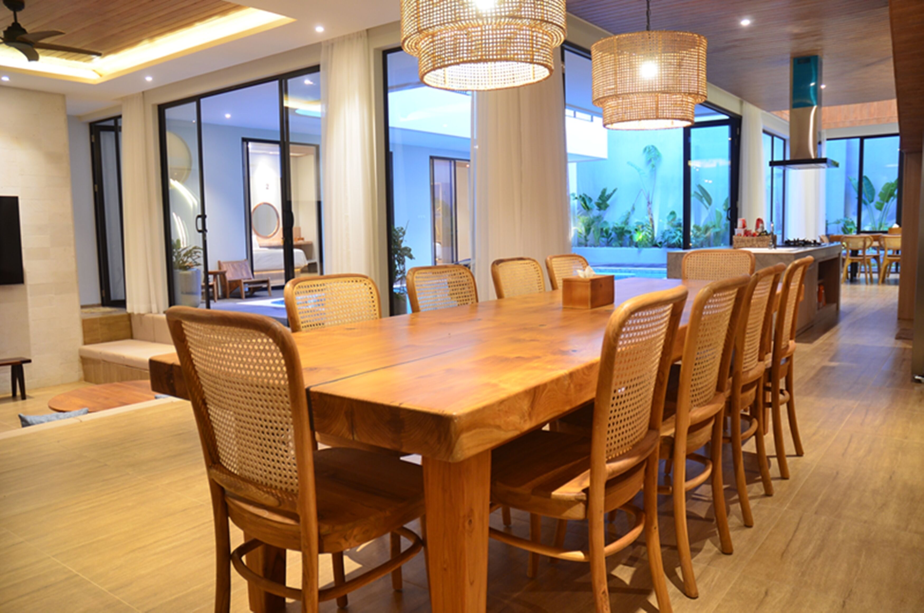 A spacious dining area with a long wooden table and wicker chairs, large windows offering a view of the outdoors, and a modern kitchen in the background.