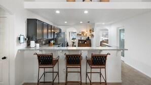 A modern kitchen with a central island, bar stools, and potted plants. - Great Location! Walk to State Farm Stadium! Heated Pool & Spa! (Glendale)