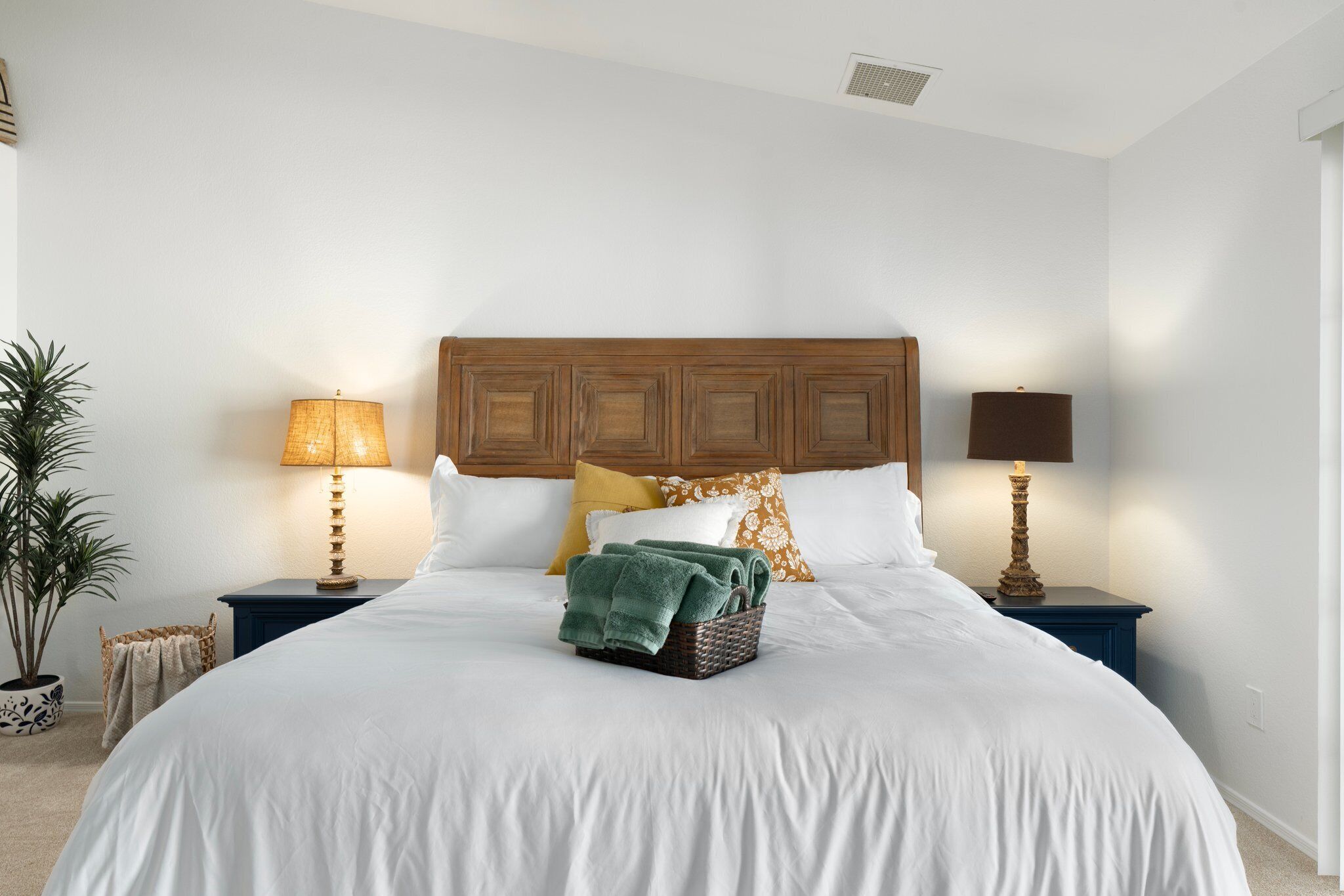 A neatly made bed with white linens, a wooden headboard, and a basket of folded towels. There are two bedside lamps and a potted plant on the left side.