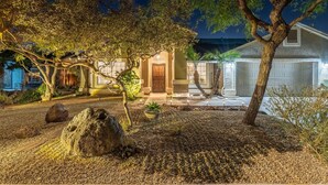 A well-lit residential property at night with a driveway, a garage, and a landscaped garden with rocks and a tree. - Great Location! Walk to State Farm Stadium! Heated Pool & Spa! (Glendale)