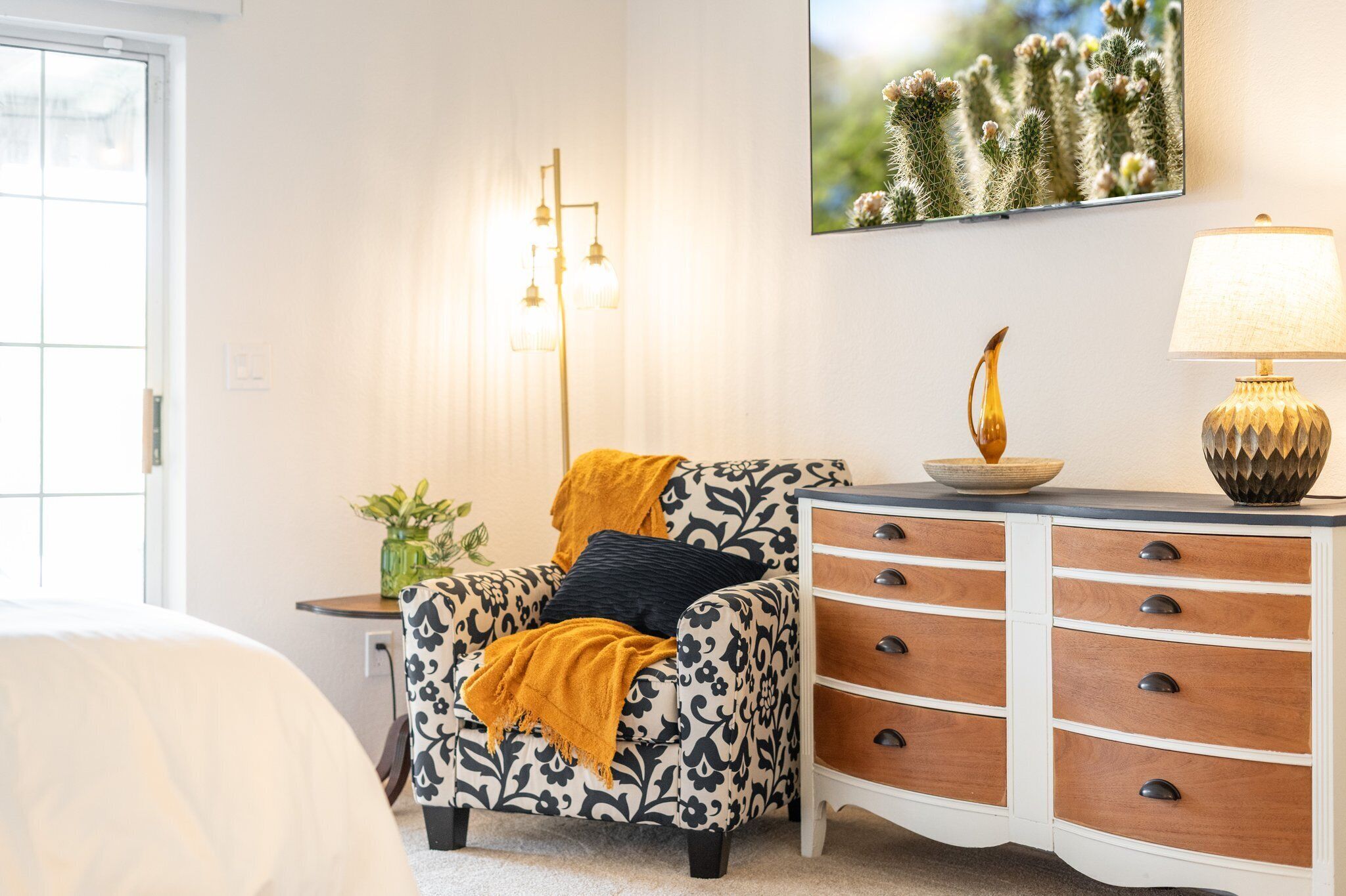 Una habitación con un sillón estampado, una mesita con una planta, una lámpara y un mueble con cajones.