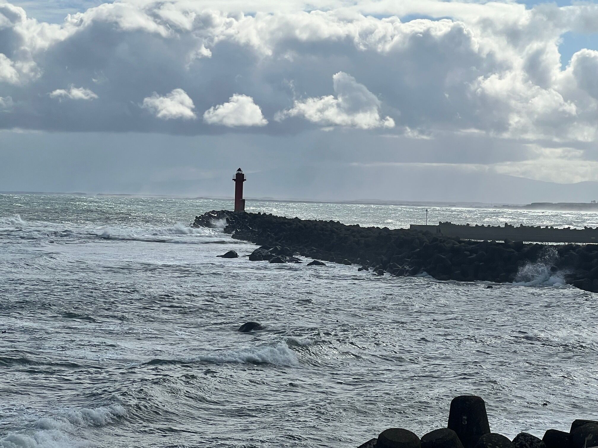 A lighthouse by the sea, waves crashing against a rocky breakwater, and a cloudy sky.