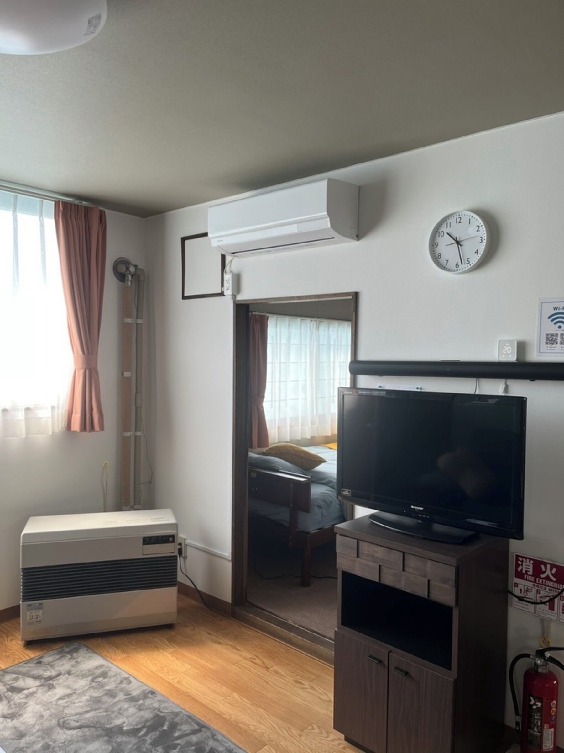 Een hotelkamer met een groot flatscreen televisietoestel, een airconditioning, een brandblusser en een klok aan de muur.