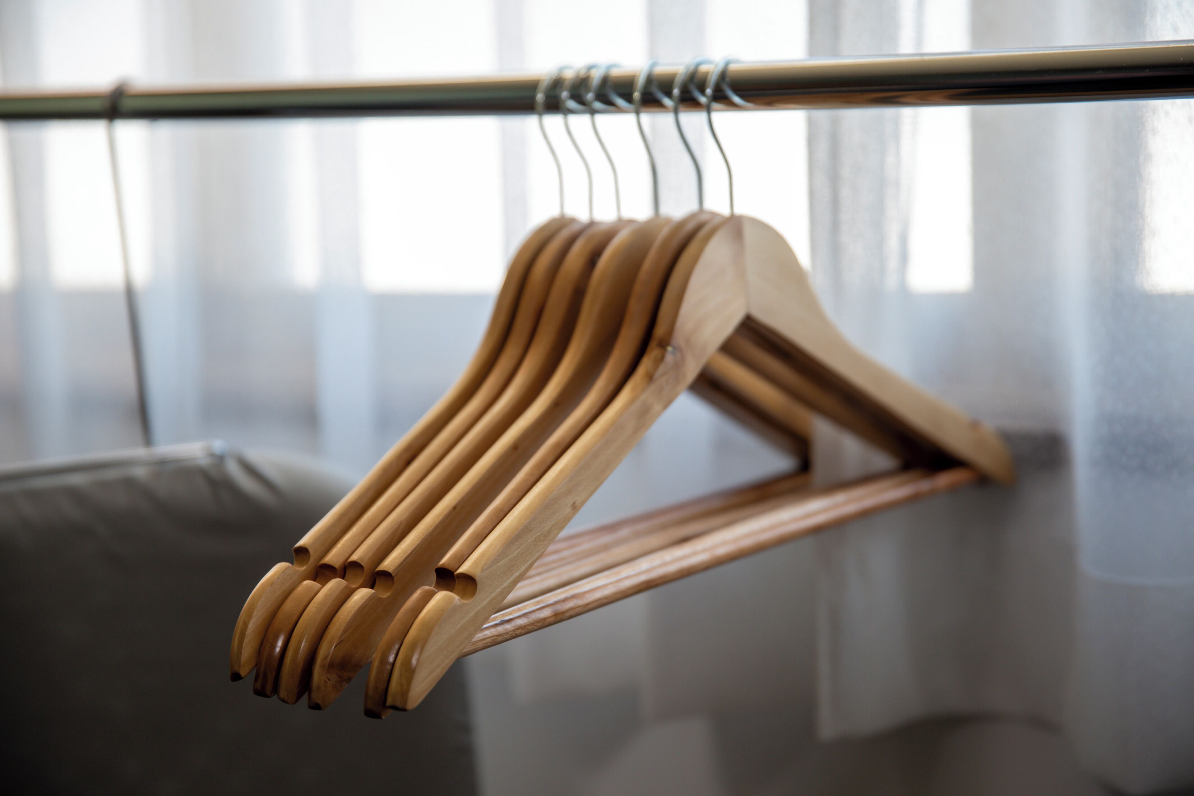 A wooden hanger on a metal rod in a room with a grey couch and a window with sheer curtains.
