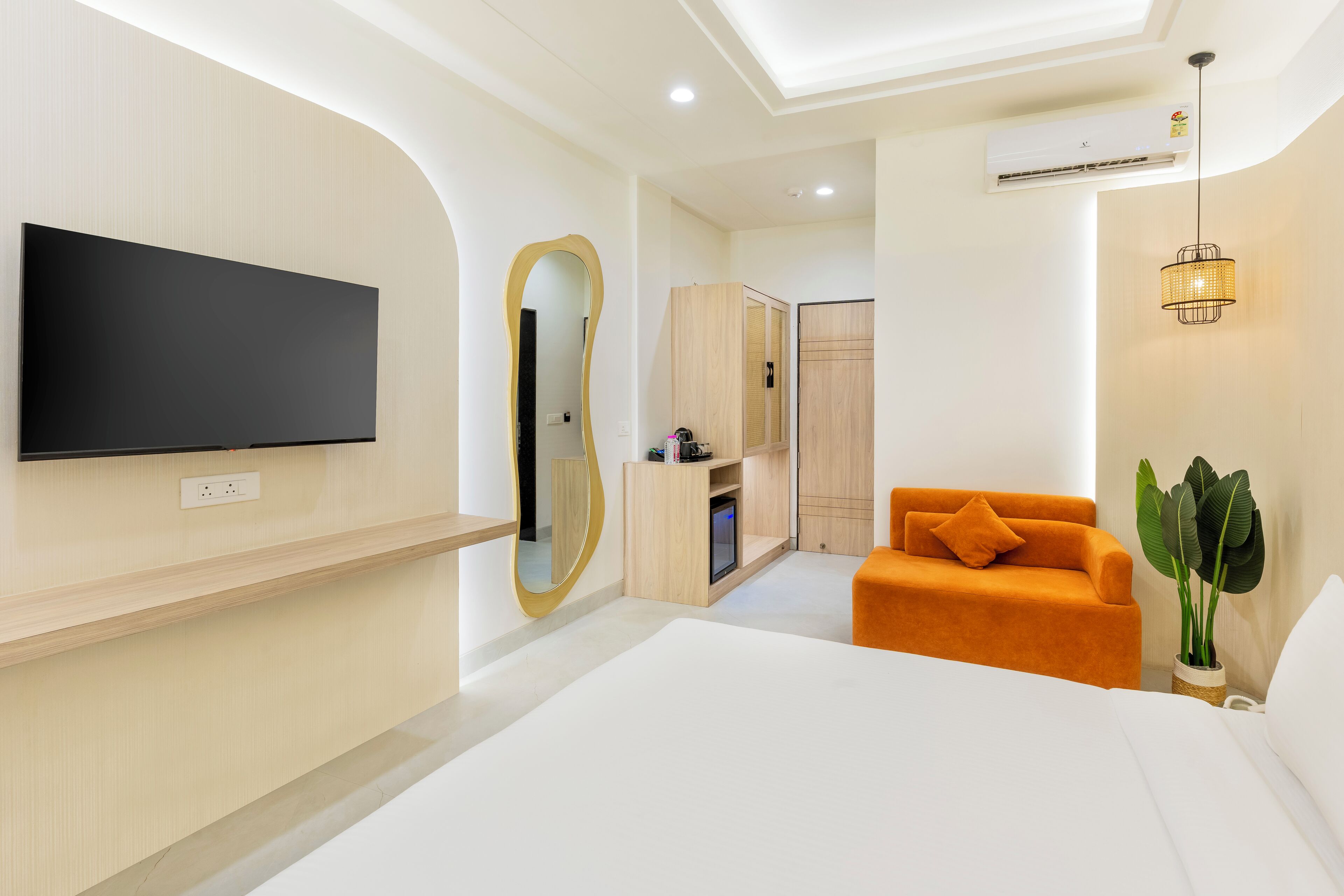 A modern hotel room with a large bed, a TV mounted on the wall, a mirror, an orange sofa, and a potted plant.