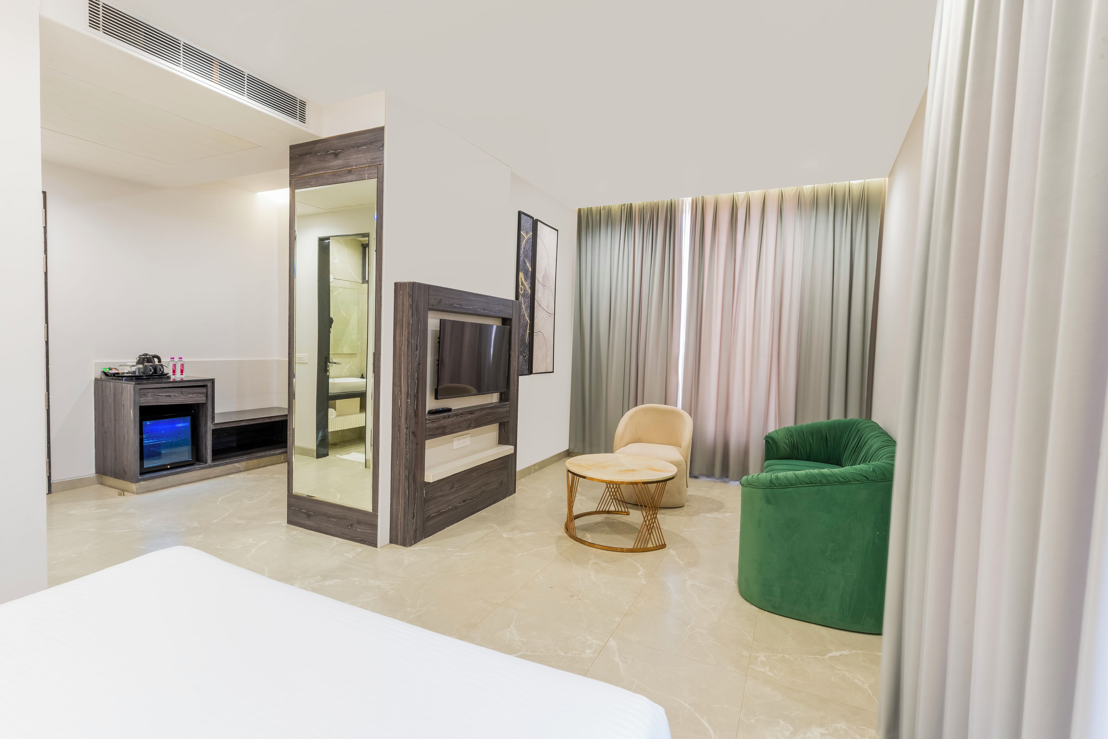 Quarto de hotel com cama, televisão, mesa pequena, cadeira verde e uma janela grande com cortinas.