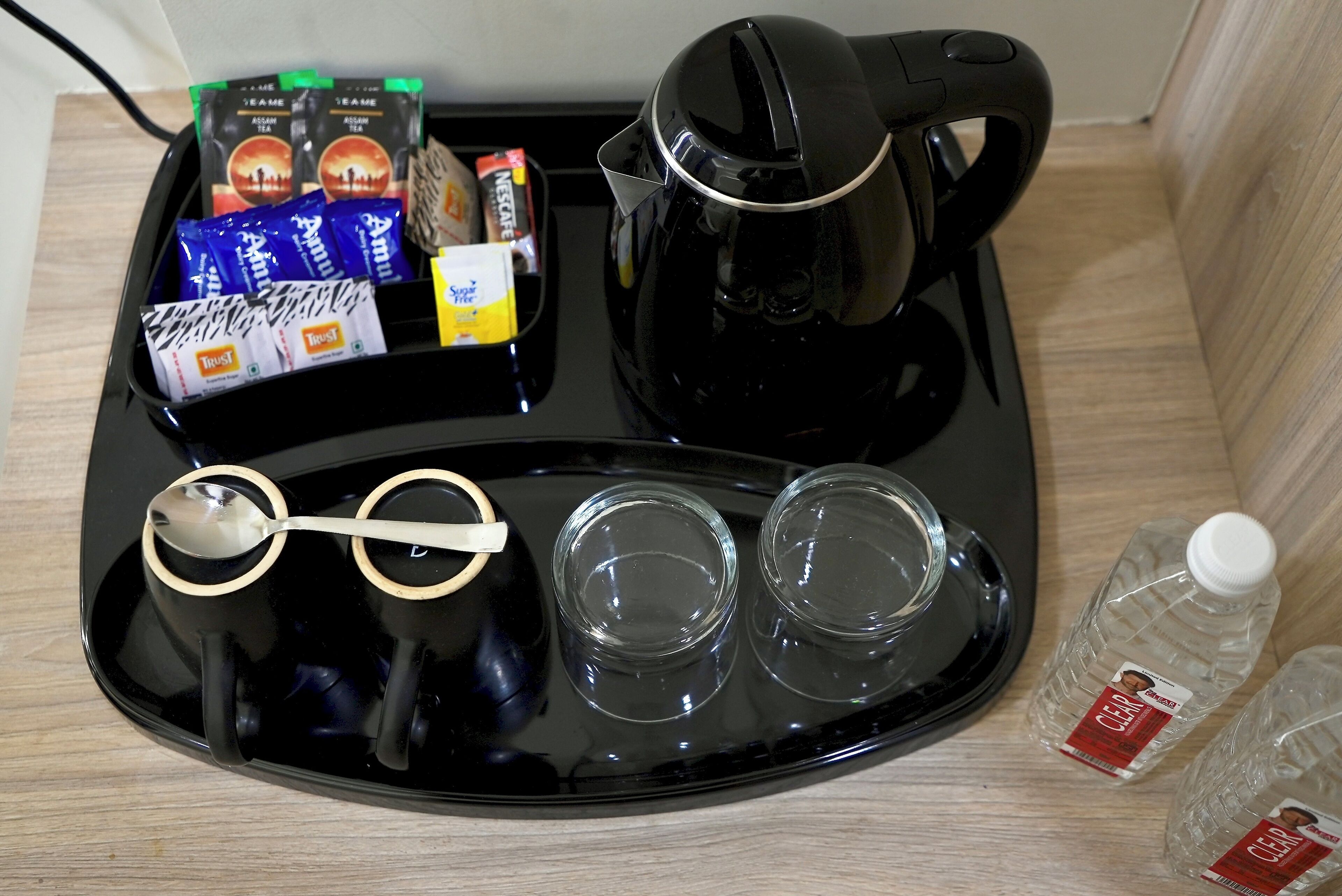 Una bandeja con una cafetera negra, dos vasos, una cuchara, bolsitas de té, una caja de azúcar y dos botellas de agua.
