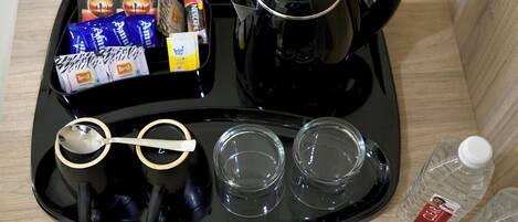 Una bandeja con una cafetera negra, dos vasos, una cuchara, bolsitas de té, una caja de azúcar y dos botellas de agua.