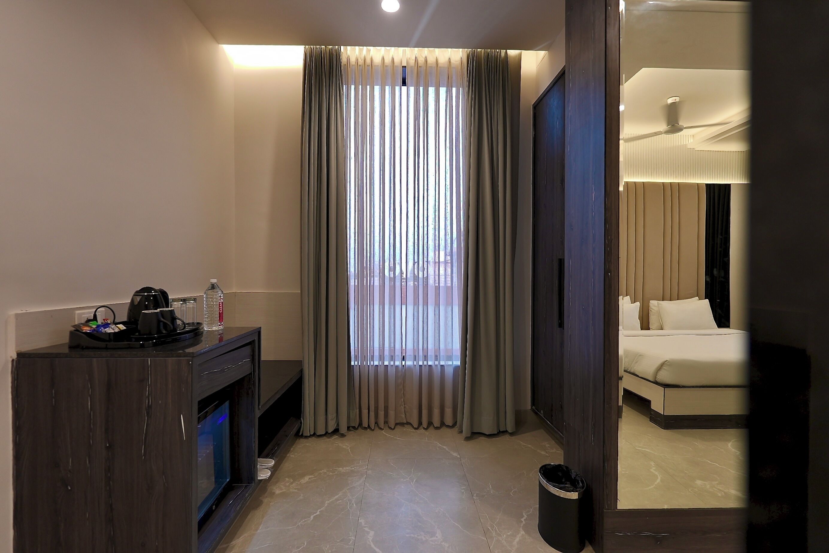 A hotel room with a bed, a desk with a coffee maker, a trash bin, and a window with curtains.