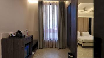 A hotel room with a bed, a desk with a coffee maker, a trash bin, and a window with curtains.