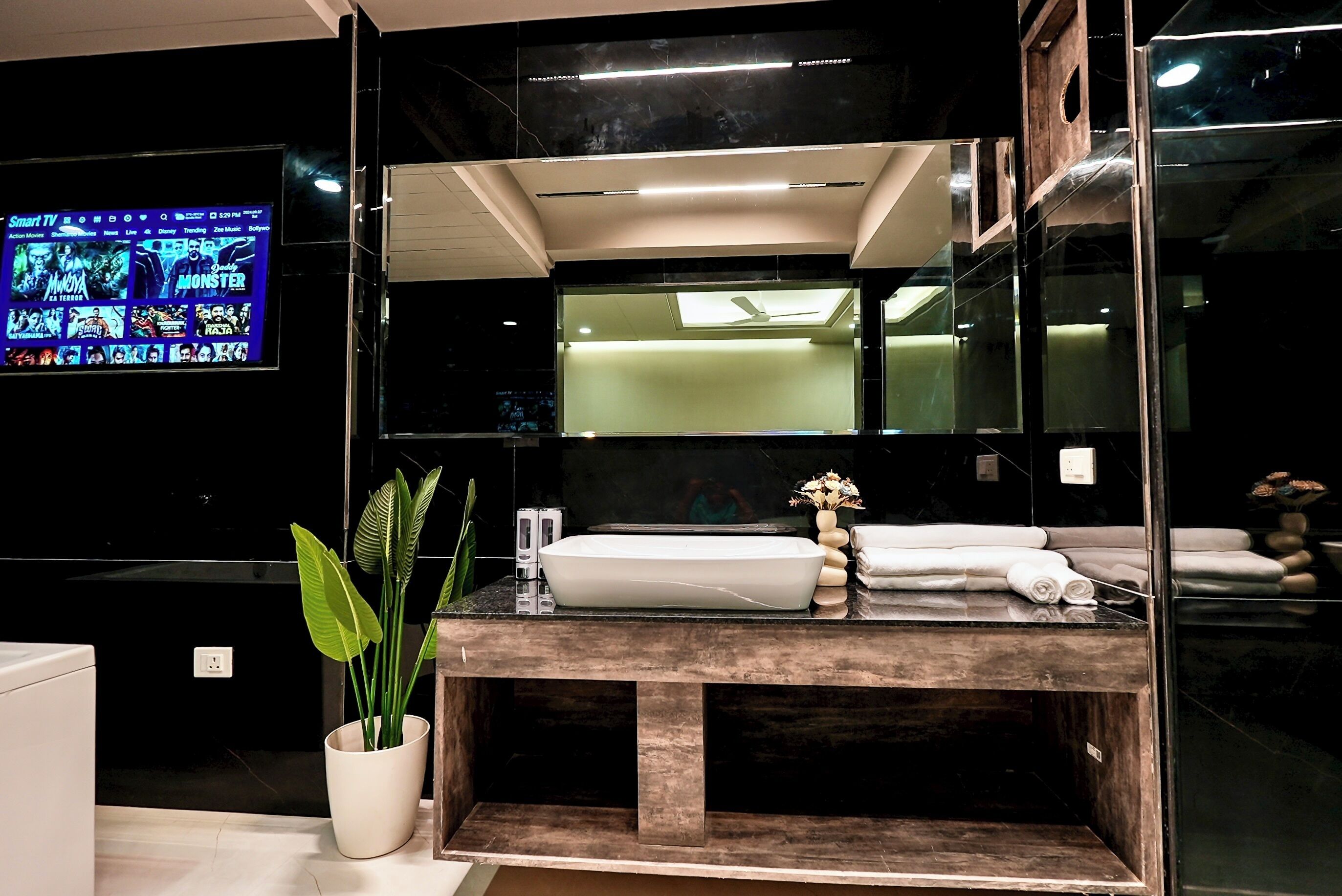 Un baño moderno con un espejo grande, un lavamanos y un televisor que muestra una selección de películas.