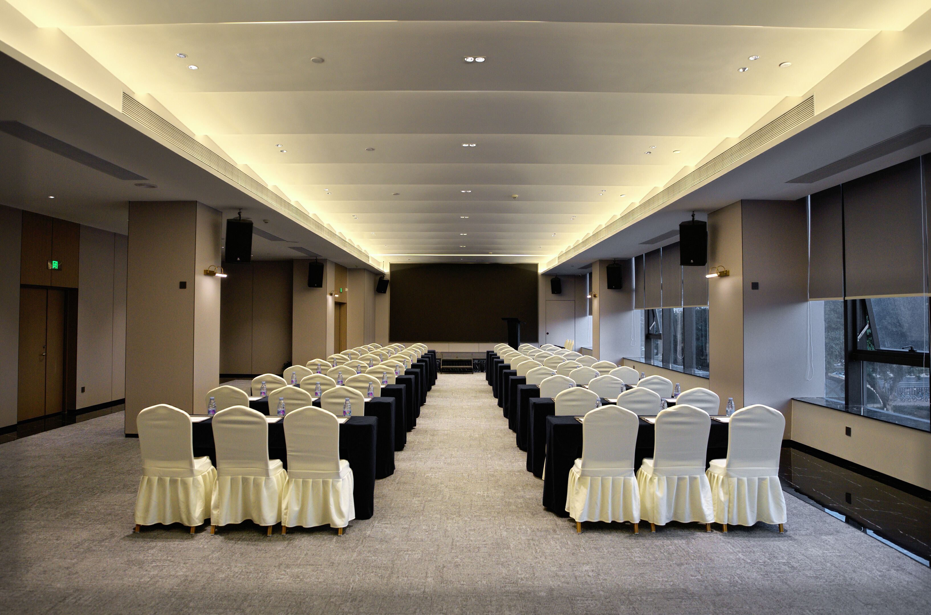 Un salón de conferencias con filas de mesas y sillas, ventanas amplias y un techo alto.