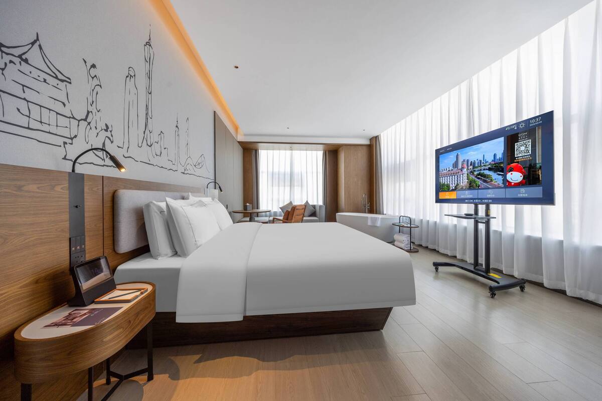Ein modernes Hotelzimmer mit einem großen Bett, einem Schreibtisch mit Laptop, einem an der Wand befestigten Fernseher und einer Stadtansicht als Wandbild.