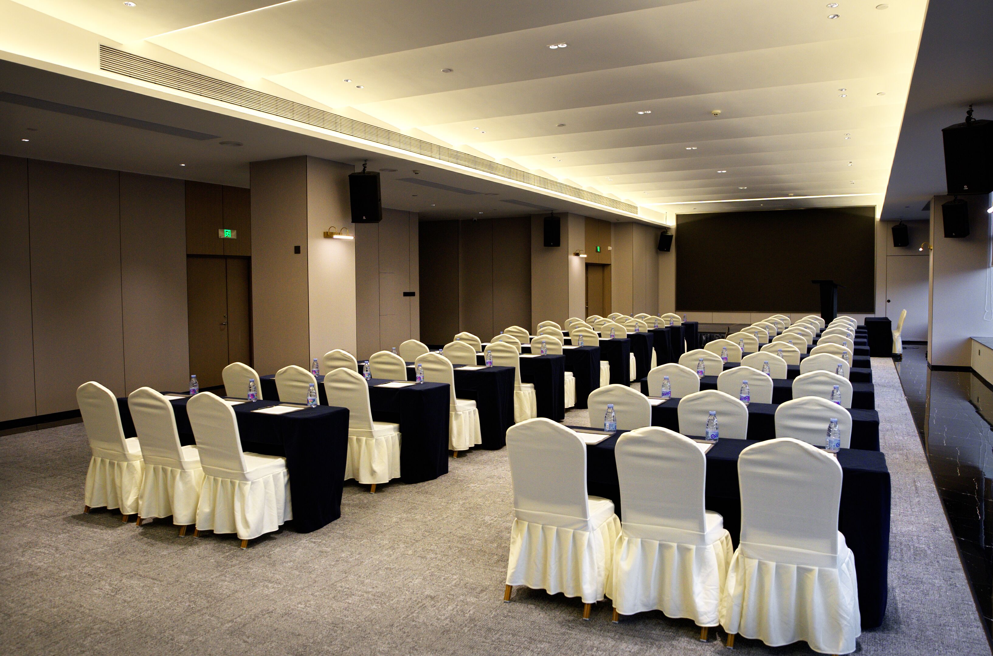 A conference room with rows of tables and chairs, equipped with water bottles, and a stage at the front.