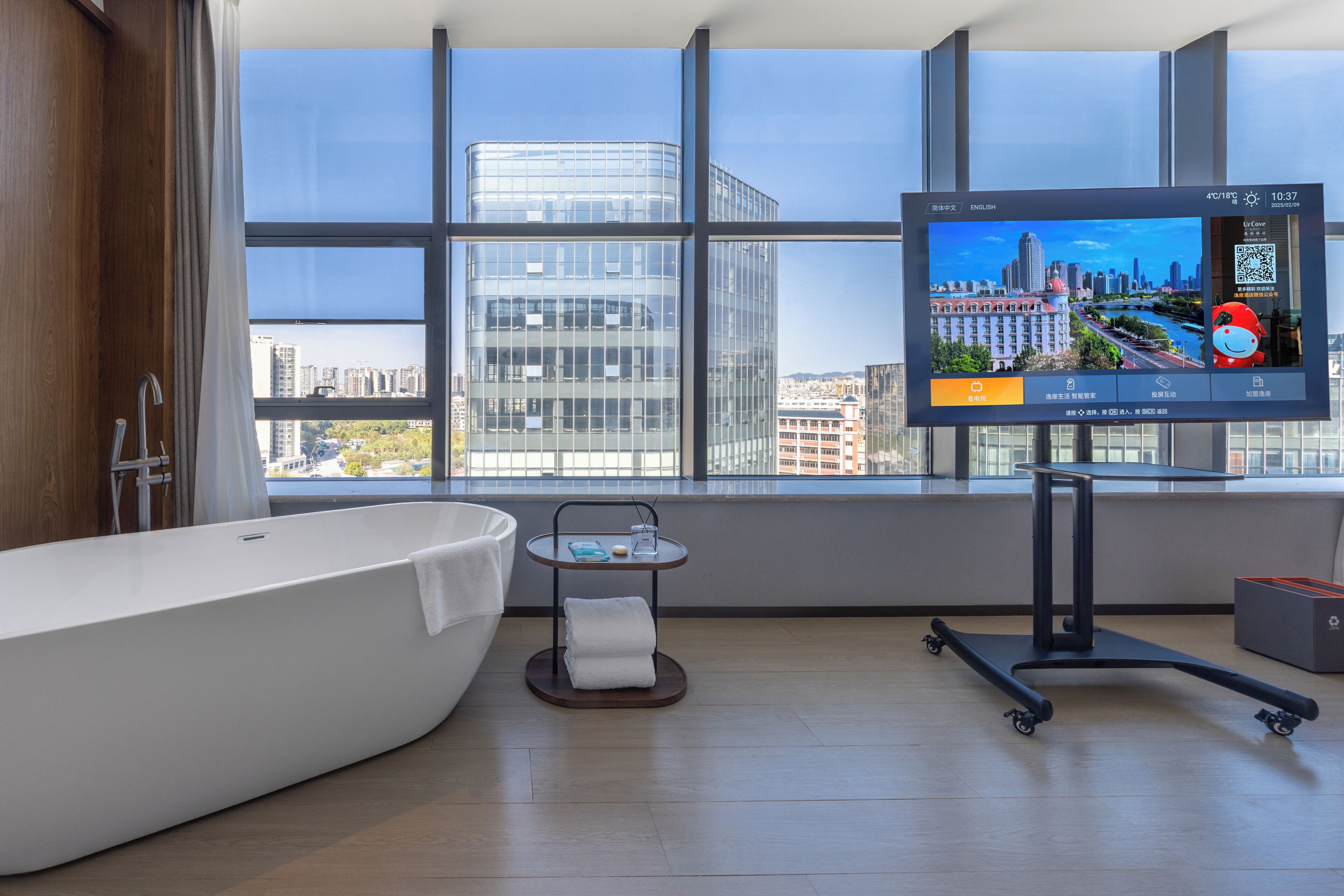 Un baño moderno con una bañera grande, un televisor montado en la pared y vistas a la ciudad a través de amplios ventanales.