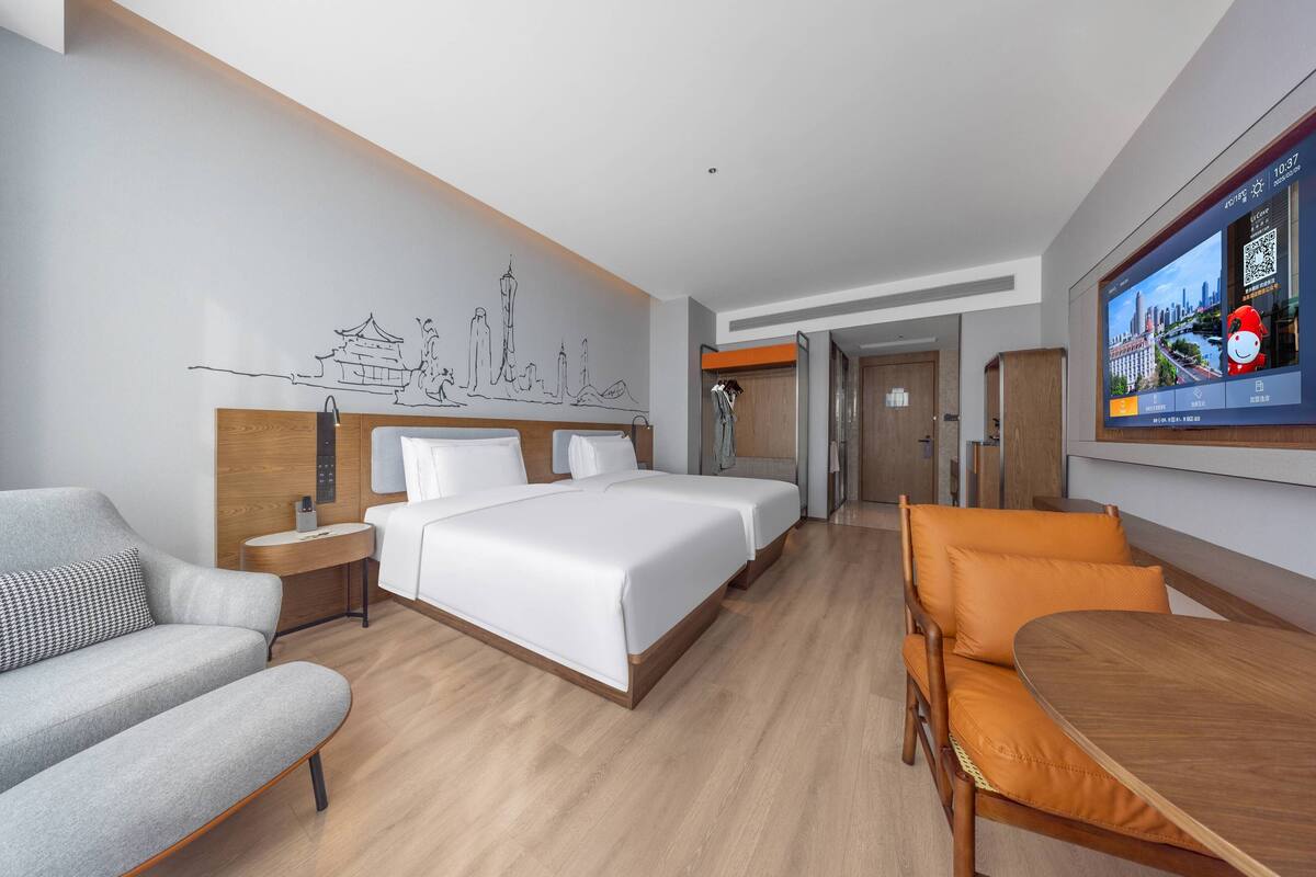 Ein modernes Hotelzimmer mit einem großen Bett, einer Sitzecke, einem Esstisch und einem an der Wand montierten Fernseher, der eine Stadtansicht zeigt.