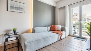 A modern living room with a grey sofa, a small side table with a plant, and a large window with a view of buildings. - Brisk - Chic 1-BR Apartment in Athens (Athens)