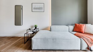 A modern bedroom with a grey sofa bed, a small bedside table with a lamp, a framed picture on the wall, and a large mirror. - Brisk - Chic 1-BR Apartment in Athens (Athens)