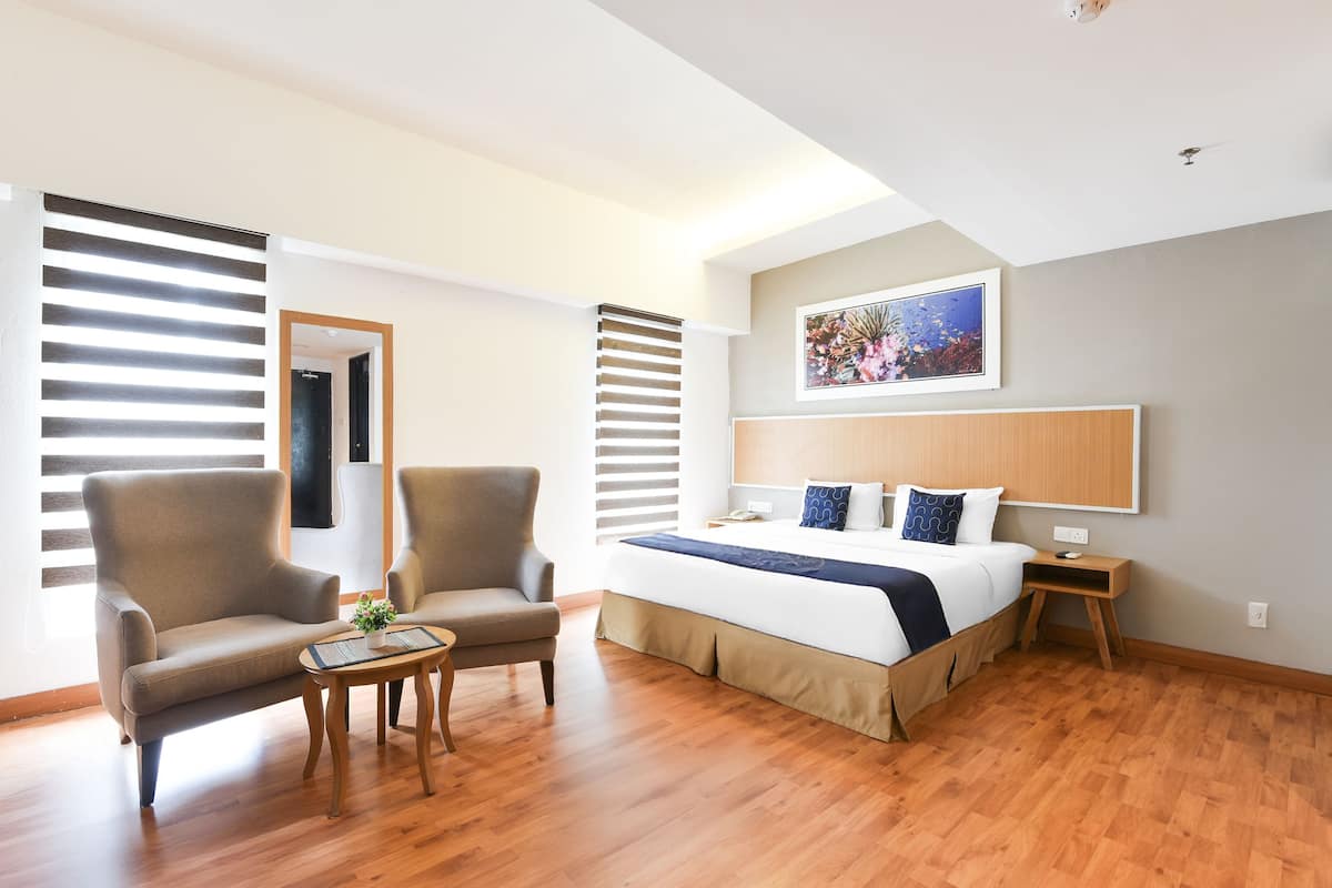 A modern hotel room with a large bed, two armchairs, a small table, and a wooden floor.