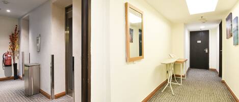 A hotel corridor with a ceiling light, a fire extinguisher, a small table, and a door at the end.