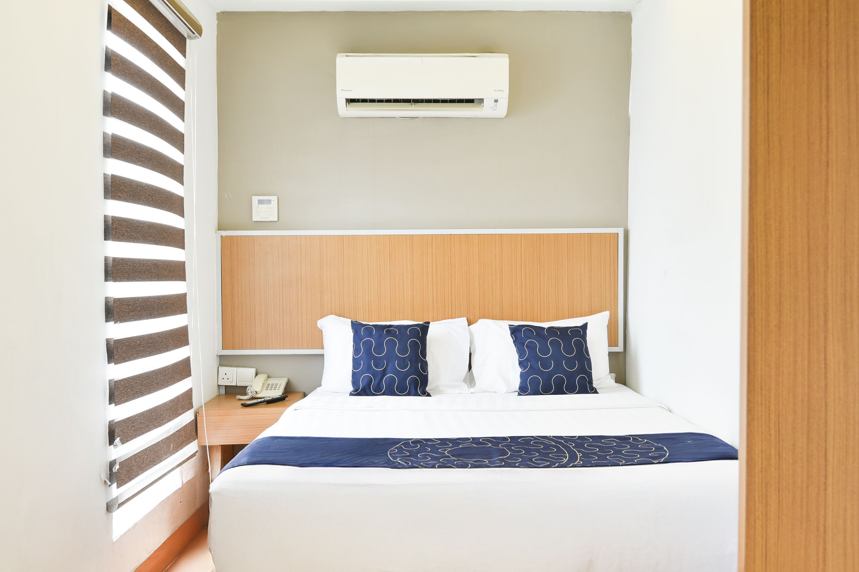 A hotel room with a bed, a bedside table with a phone, a wall-mounted air conditioner, and a window with blinds.