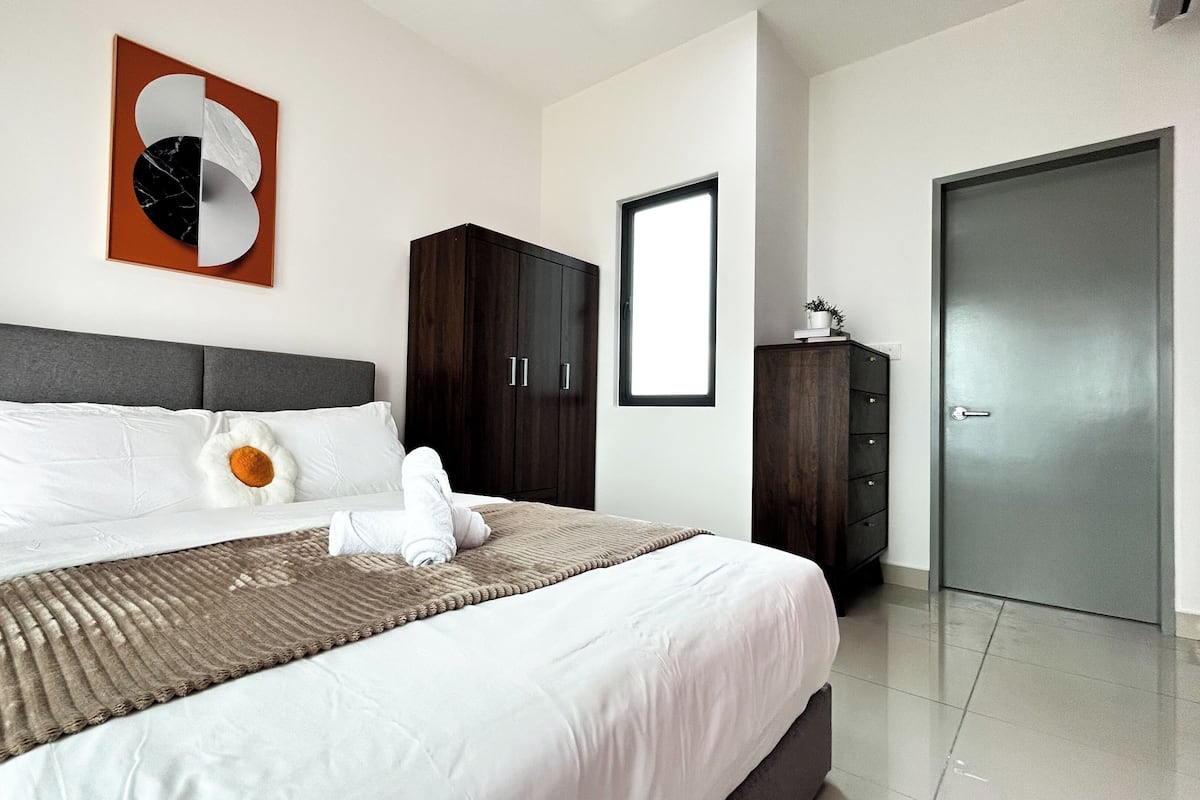 Uma cama bem arrumada com lençóis brancos e um cobertor marrom, um guarda-roupa de madeira escura e uma porta cinza.