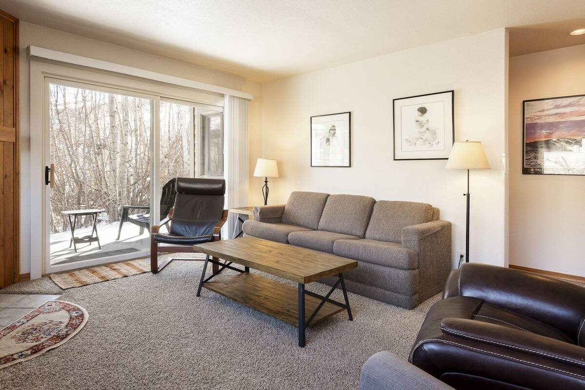 Sala de estar com sofá, poltrona, mesa de centro e abajur. Quadros emoldurados na parede e janela com vista para árvores.