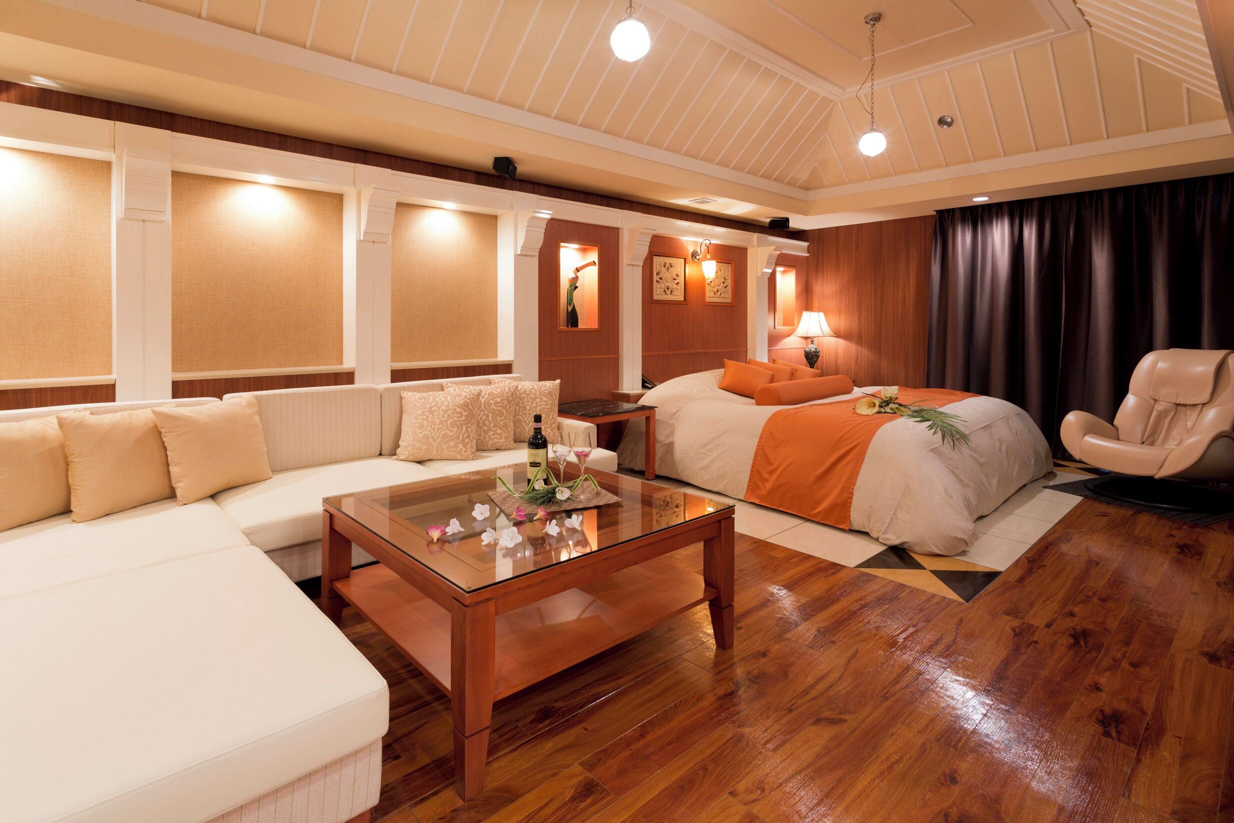 Une chambre d’hôtel avec un plancher en bois, un canapé, un lit recouvert d’une couverture orange, une chaise et une table basse en verre.