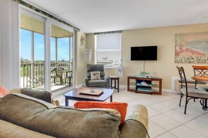 Un salon comprenant un canapé, un fauteuil et une table à manger avec des chaises. Un téléviseur est fixé au mur et il y a un balcon avec vue sur l’extérieur.