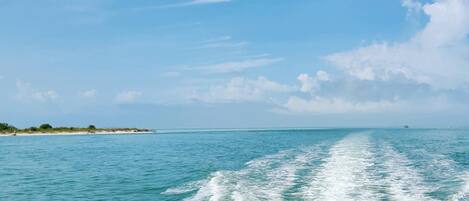 A clear view of the ocean with a wake behind a moving boat, a distant shoreline with vegetation, and a partly cloudy sky.