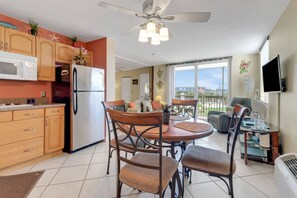 Cocina compacta con refrigerador, microondas y armarios de madera. Cuenta con un comedor con mesa y sillas, y un balcón con vistas a la ciudad.
