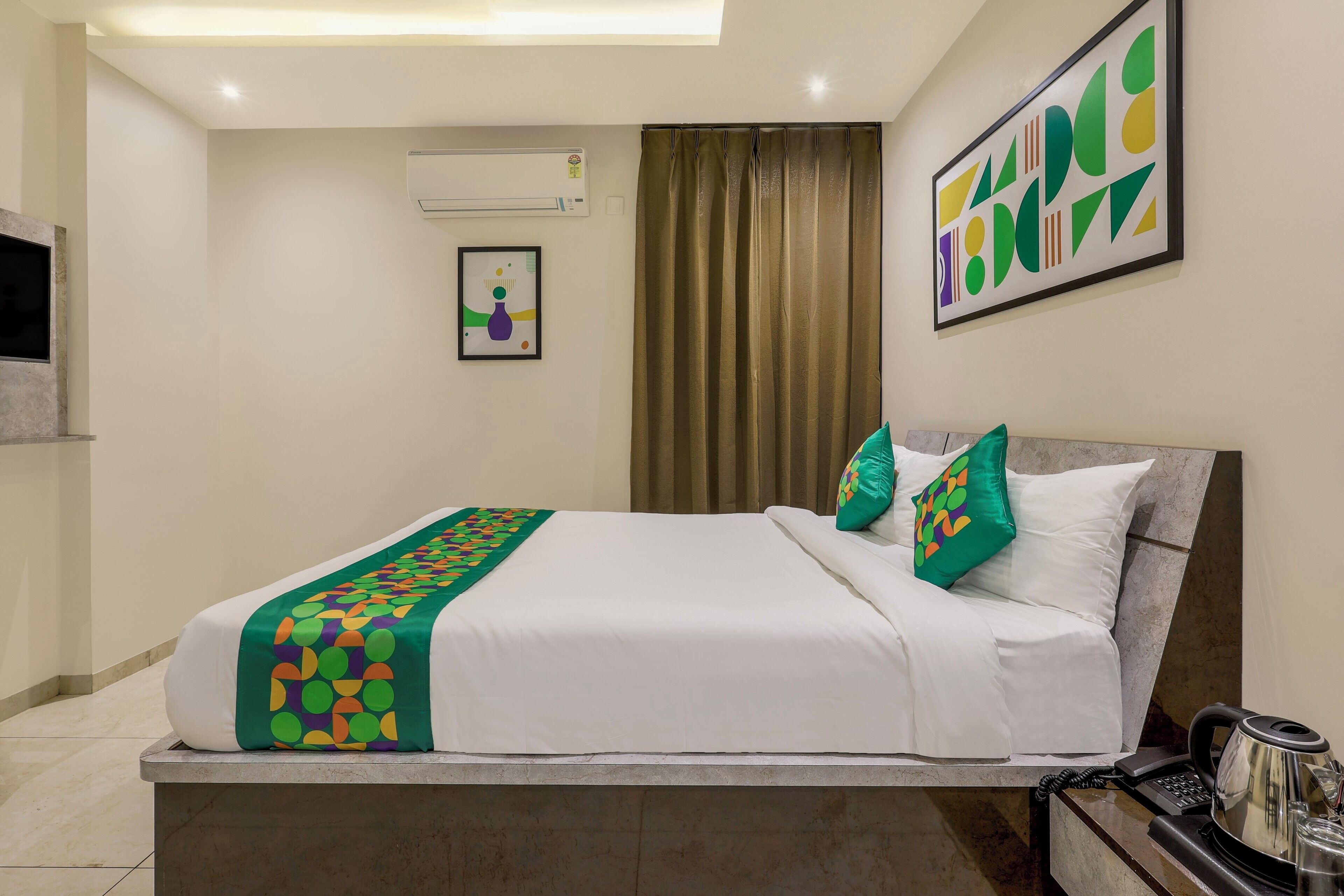 A hotel room with a bed, a bedside table with a telephone and a kettle, a wall-mounted air conditioner, a framed abstract painting, and a window with curtains.