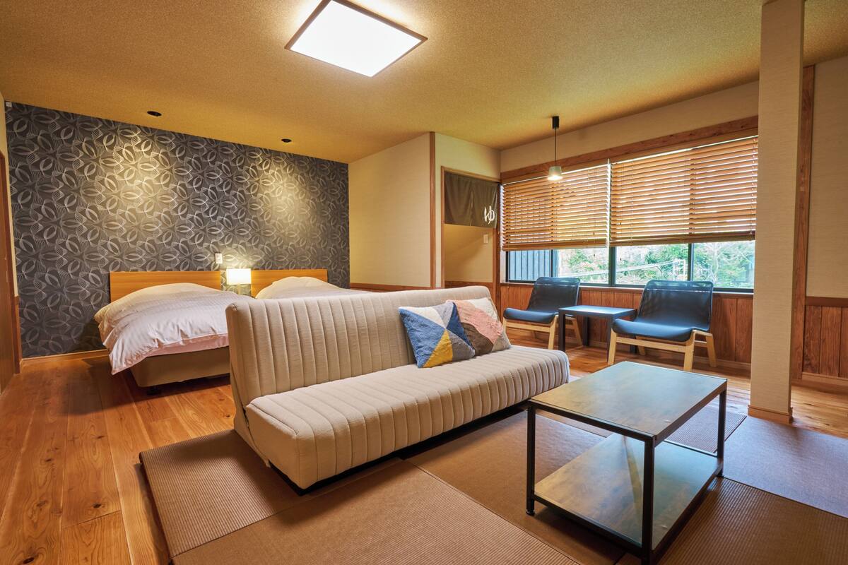 Een kamer met een bed, een bank, een klein tafeltje en een raam met jaloezieën.