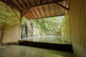 Un onsen japonais traditionnel, doté d’un toit en bois et d’un sol carrelé, entouré de formations rocheuses naturelles et de verdure.