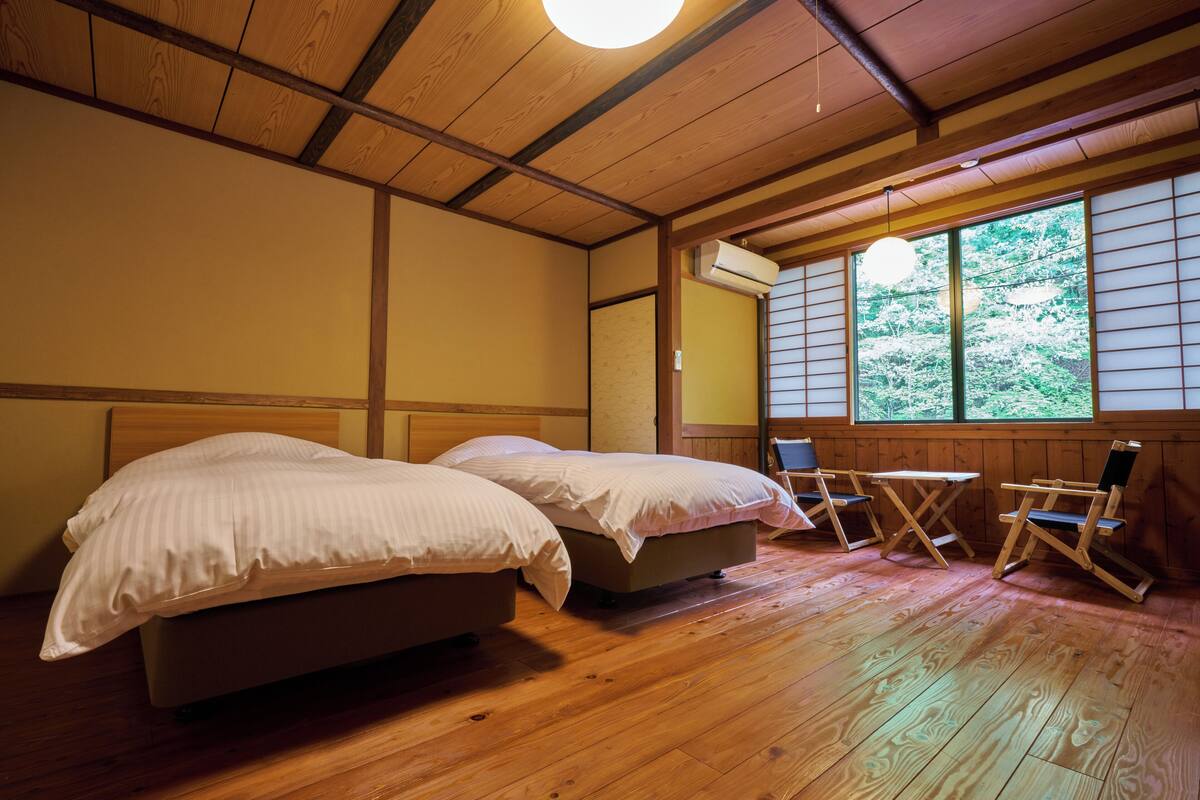 Een kamer met twee bedden, een houten vloer, een raam met uitzicht op bomen en een bureau met stoel.