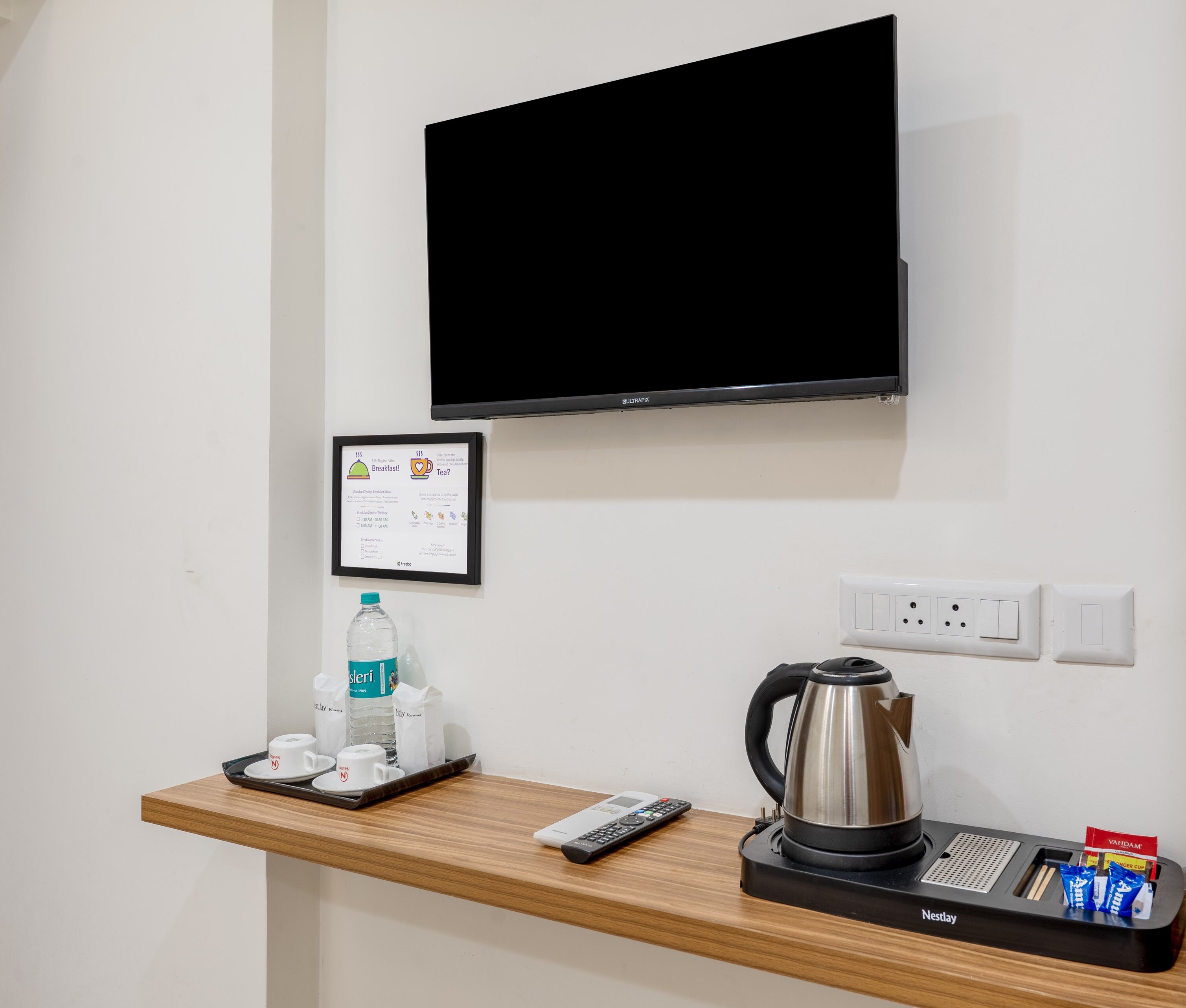 Ein Hotelzimmer mit einem wandmontierten Flachbildfernseher, einer kleinen Holztrennwand mit einem Wasserkocher, einer Fernbedienung, einer Ablage mit Tassen und einer Wasserflasche sowie einer gerahmten Speisekarte an der Wand.