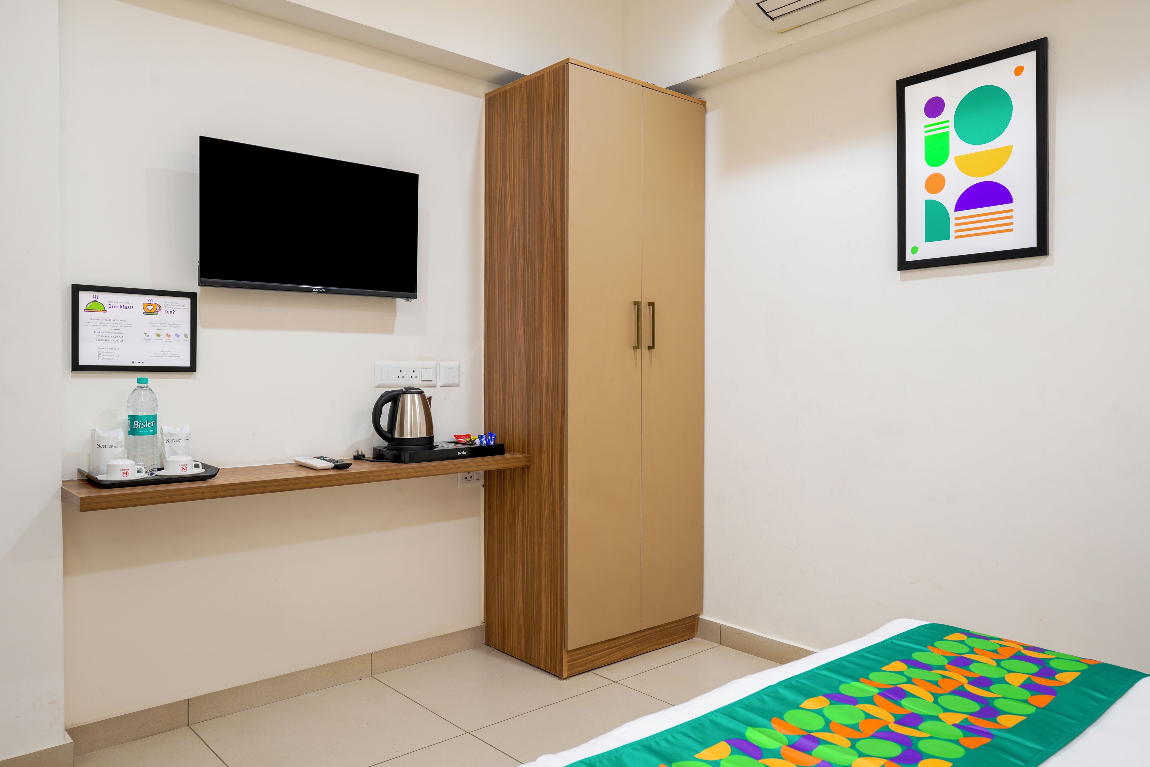 A room with a bed, a wooden wardrobe, a mounted TV, a framed abstract art piece, and a small table with a kettle and a bottle.