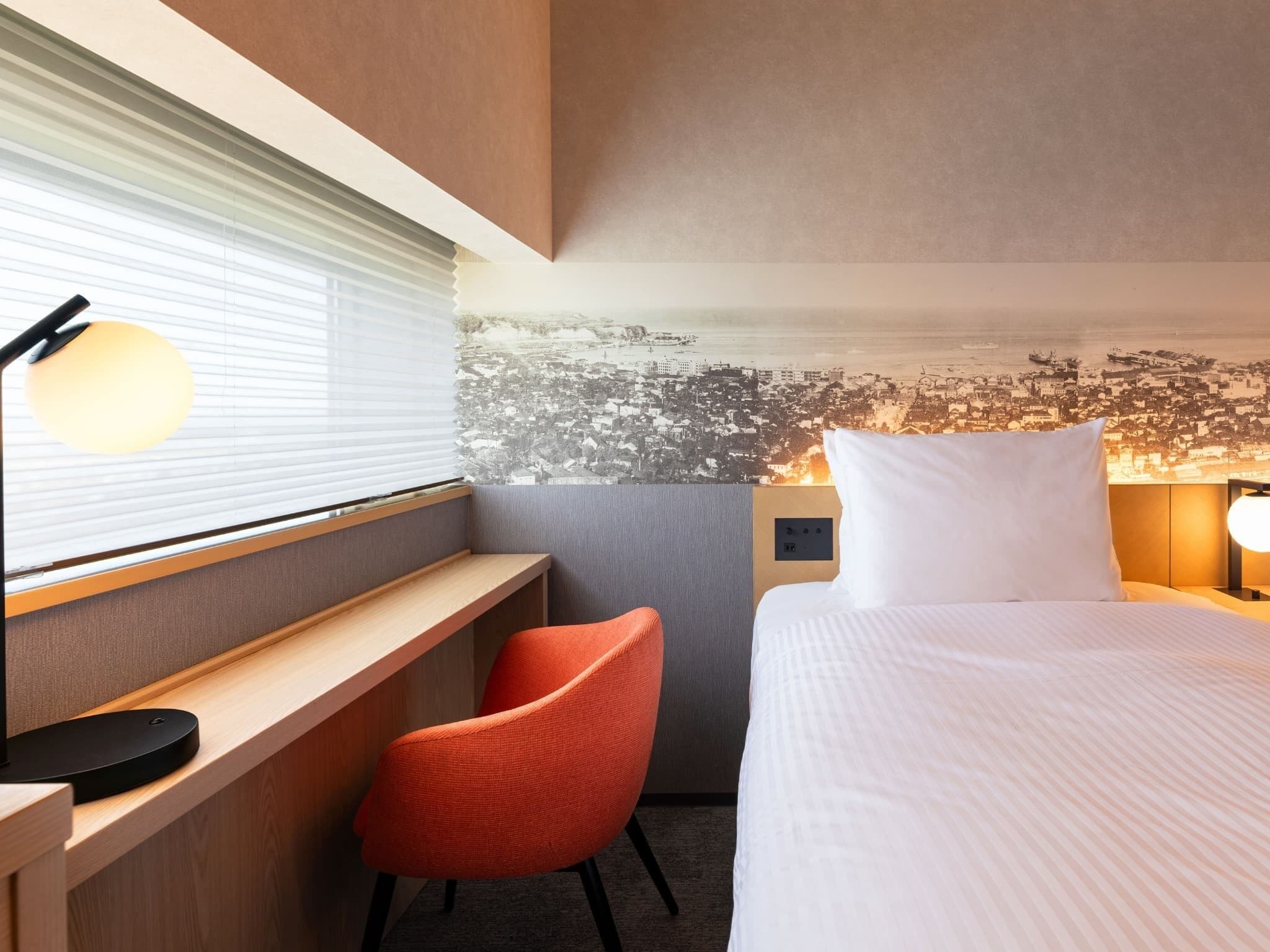 Habitación de hotel con cama, escritorio, silla, ventana con persianas y un mural en la pared que representa un paisaje urbano.
