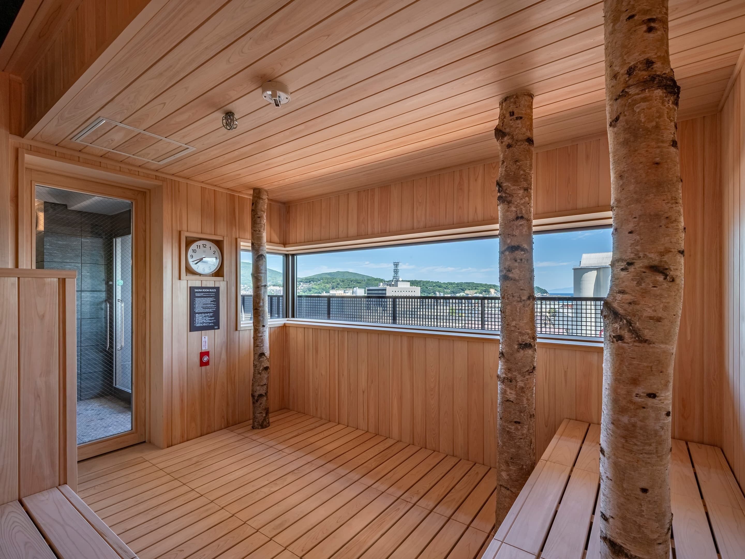 A sauna room with wooden walls, a wooden floor, a clock, and a window with a view of a cityscape.
