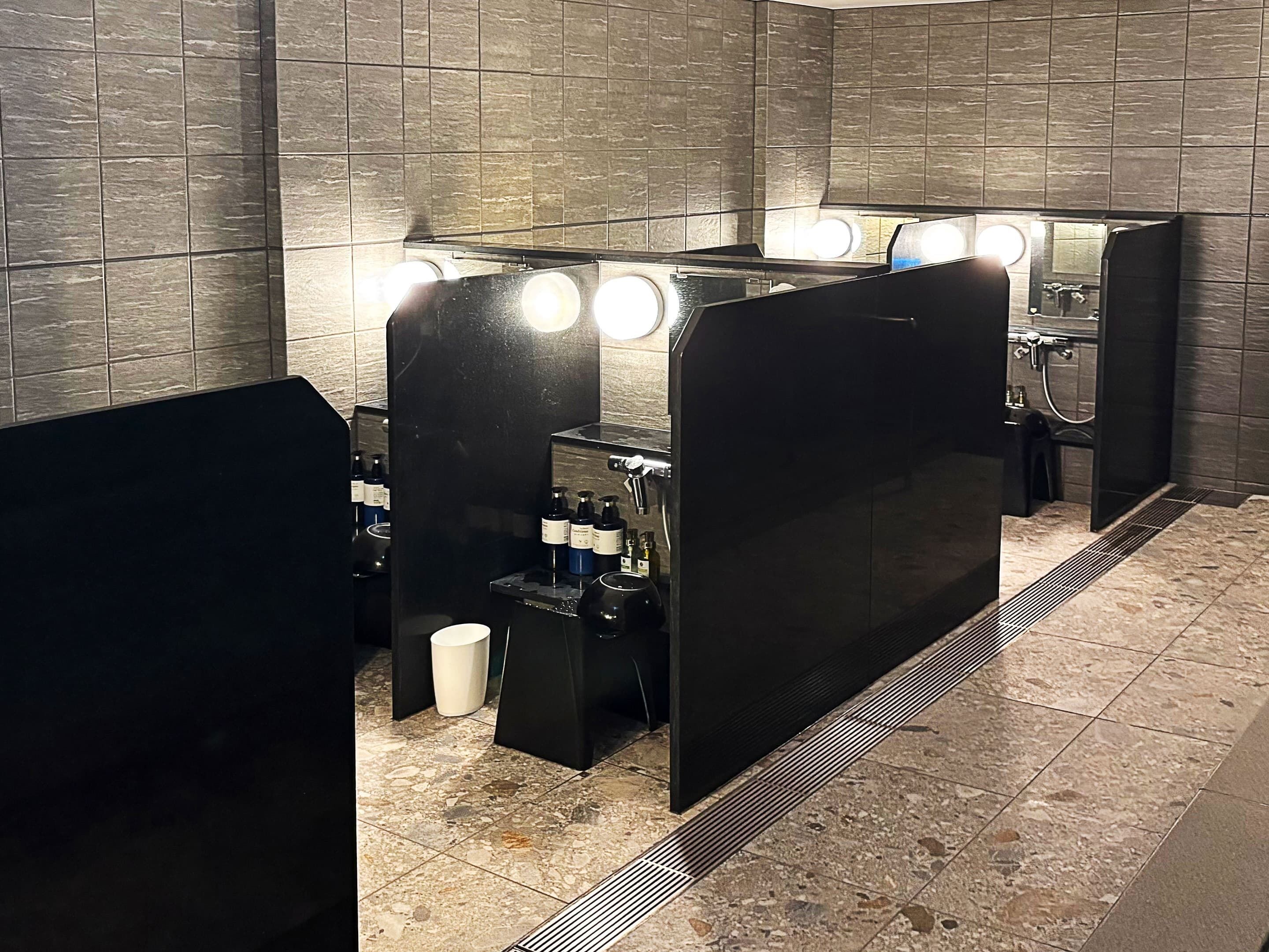 A modern bathroom with dark tiled walls, a black vanity with a mirror and lights, and a shower area.