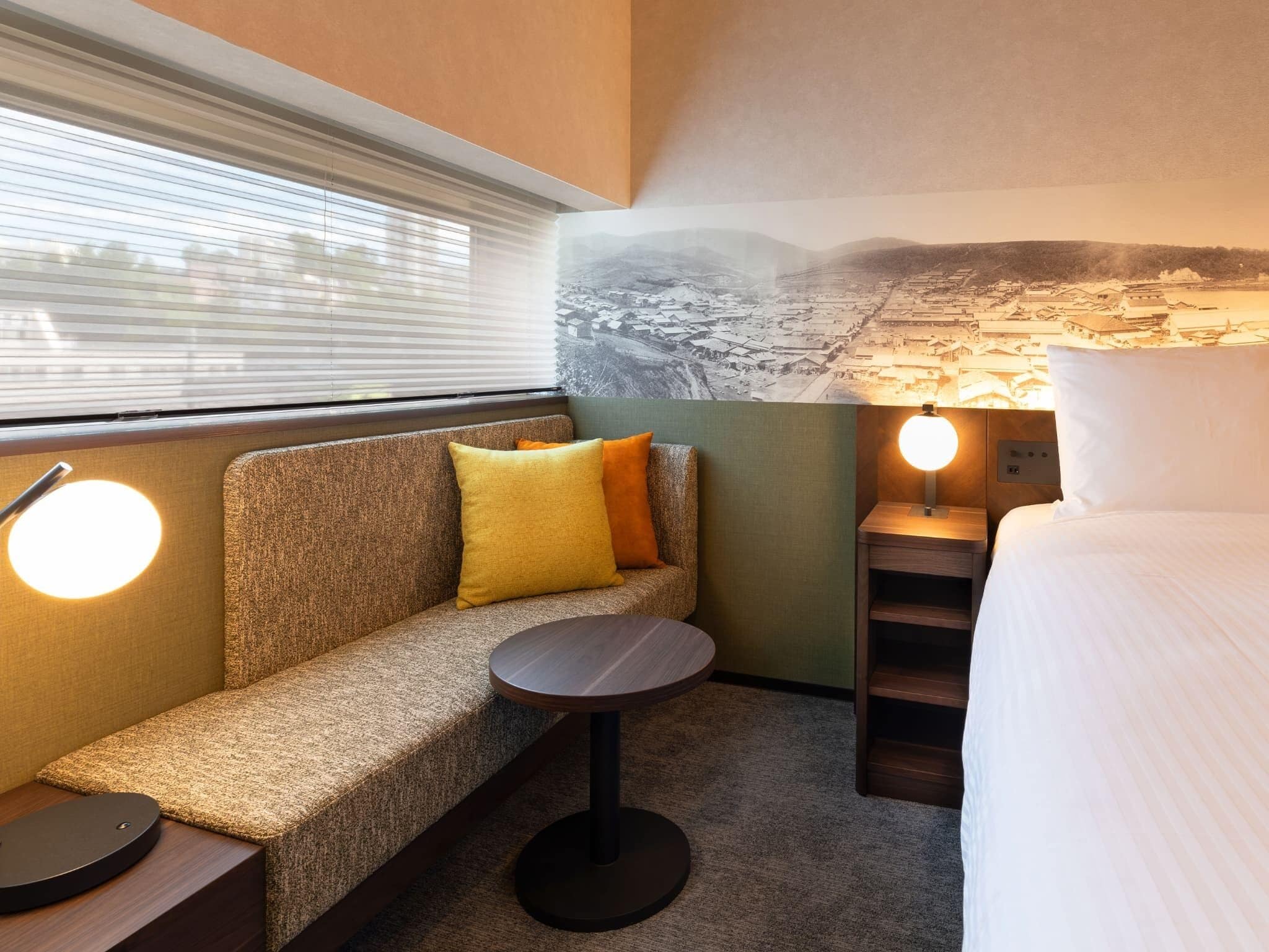 A compact hotel room with a sofa, a small round table, a bed, and a wall-mounted lamp.