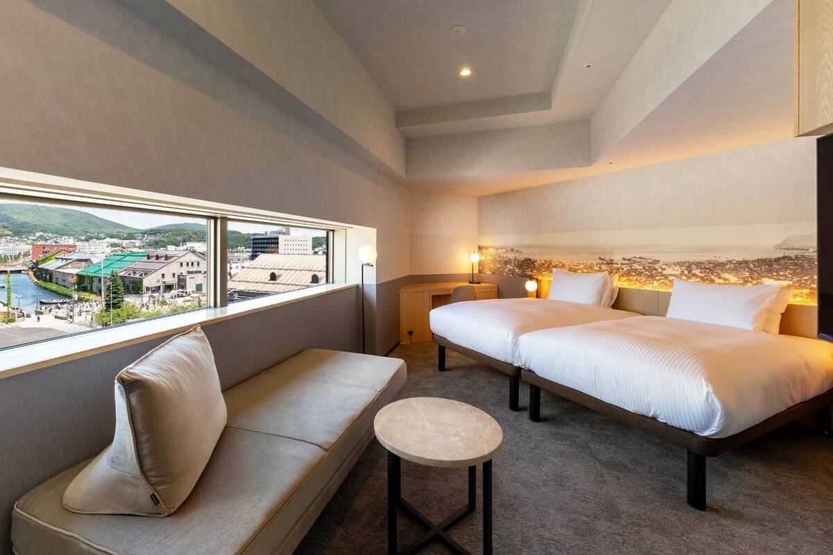 A hotel room with a bed, a sofa, a small table, and a view of a cityscape through a large window.
