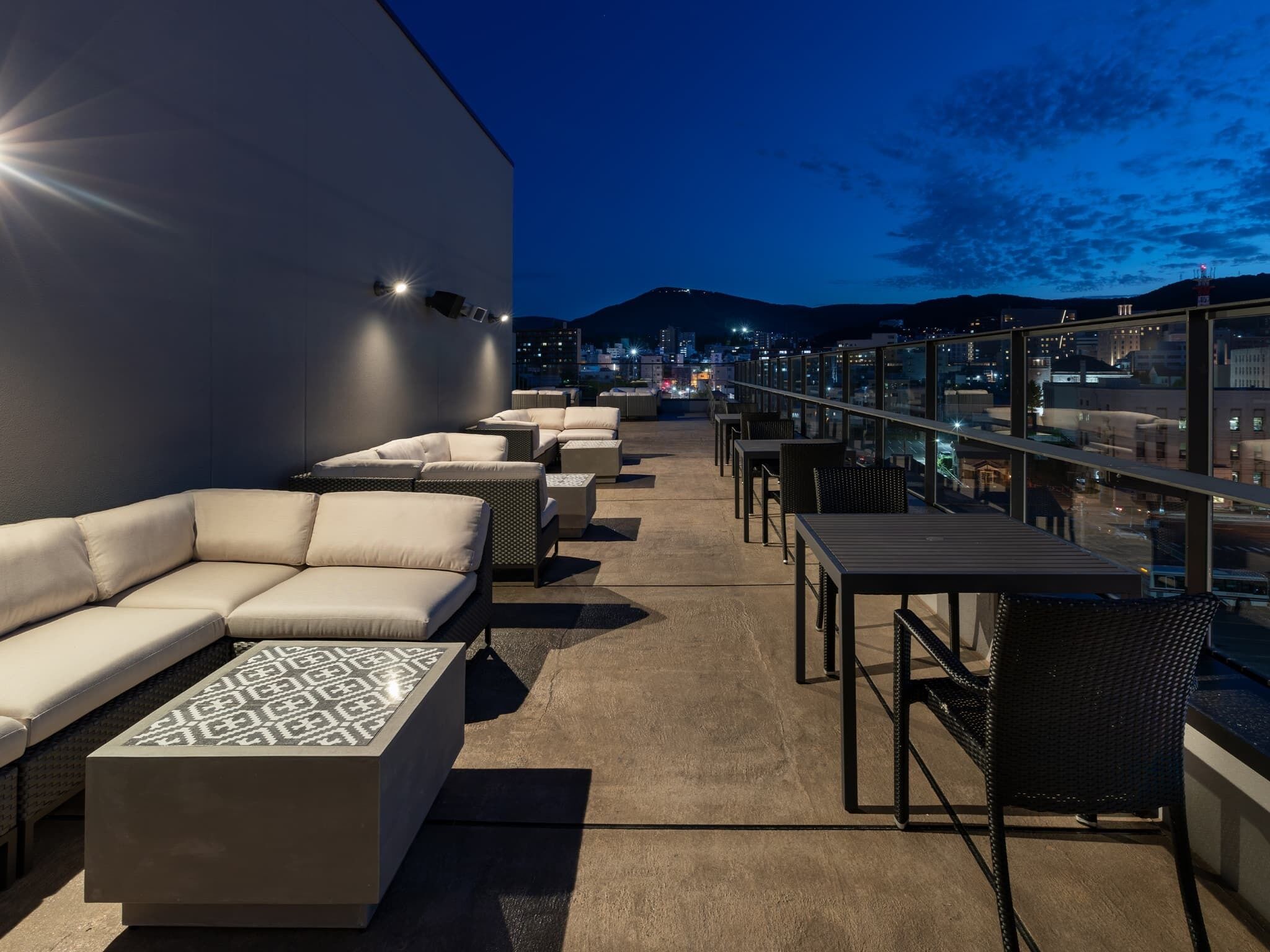 Terraza en la azotea con sofá, mesas y sillones, con vistas a un paisaje urbano al atardecer.