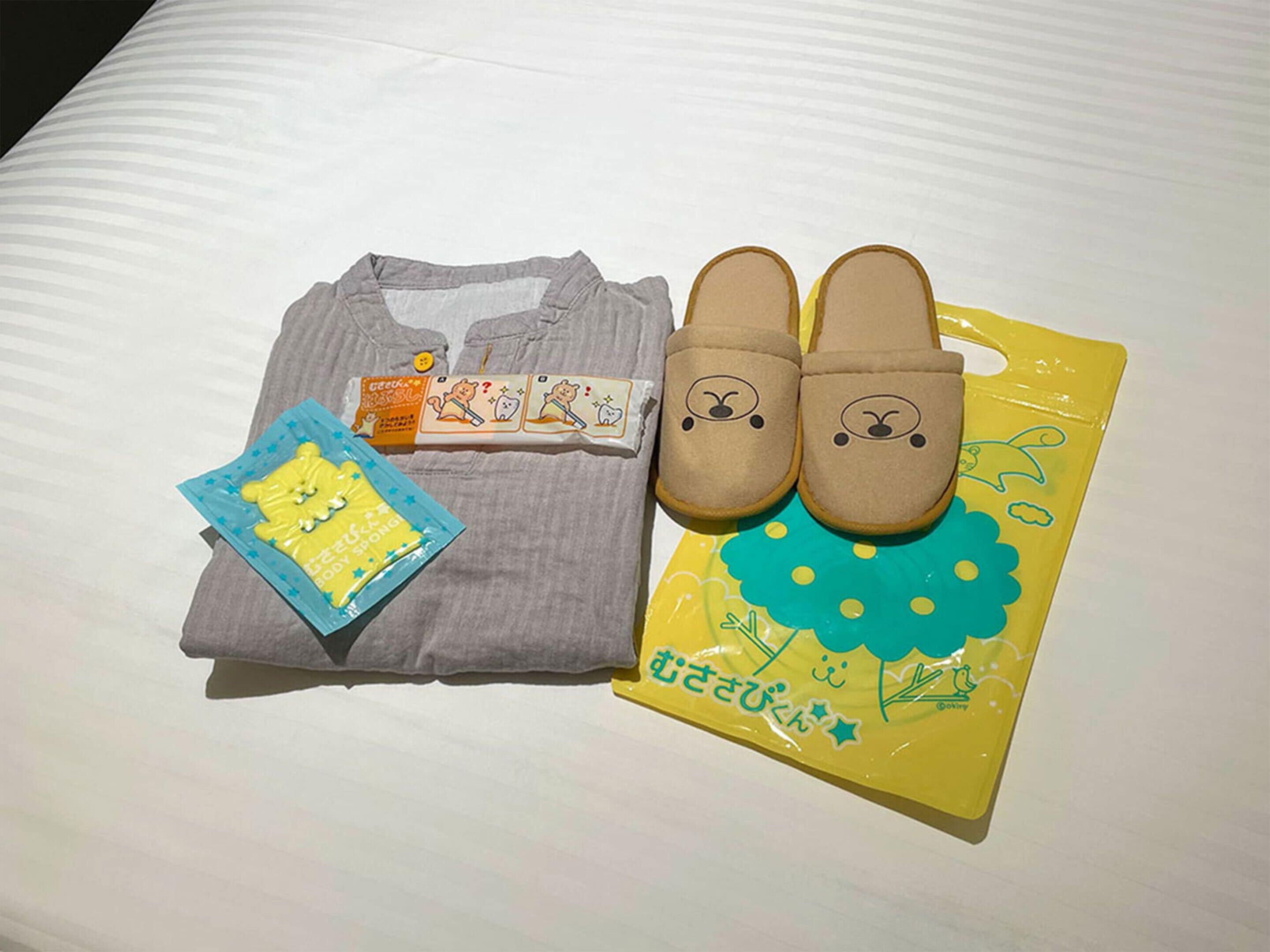 A neatly folded gray t-shirt, a pair of brown slippers with a bear face design, a yellow bag with a bear and flowers, and a small packet of yellow sponge.