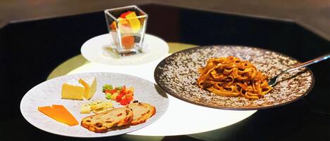 A dining table with a plate of pasta, a plate with cheese and bread, and a small glass of dessert.
