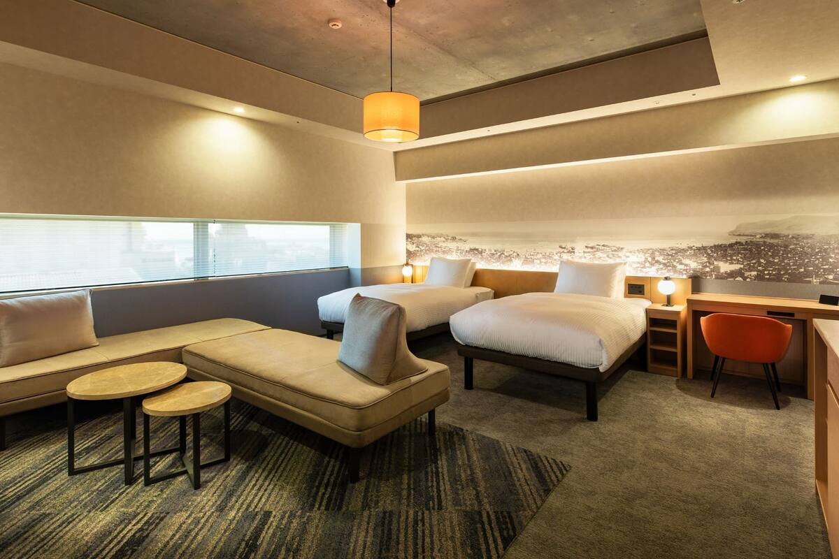 A modern hotel room with a sofa, two beds, a desk, and a cityscape mural on the wall.